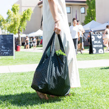 Load image into Gallery viewer, At an outdoor market, a person in a white dress carries a Bumkins Packable Tote Bag: Rich Black with greenery peeking out.

