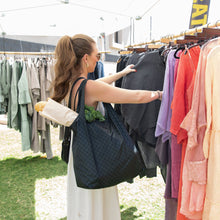 Load image into Gallery viewer, A woman shops at an outdoor market, carrying a Bumkins Packable Tote Bag: Rich Black with a baguette and greens.
