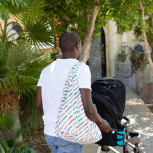 Load image into Gallery viewer, Someone in a white shirt walks with a Bumkins Packable Tote Bag: Mickey &amp; Friends Varsity on a sunny path, pushing a stroller.
