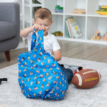 Load image into Gallery viewer, A young boy sits with his Bumkins Packable Tote Bag: Mickey &amp; Friends Forever, next to a football against toy-filled shelves.

