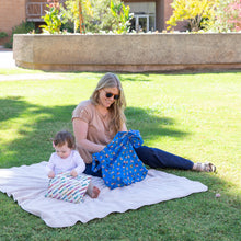 Load image into Gallery viewer, In the park, a woman holds a Bumkins Packable Tote Bag: Mickey &amp; Friends Forever near her toddler playing with a colorful bag on a blanket.

