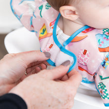 Load image into Gallery viewer, Adult hands fasten the hook-and-loop closure of a Bumkins Ruffle Apron Bib: Boots around a child's neck.
