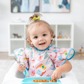 A toddler sits in a high chair, wearing a Bumkins Ruffle Apron Bib: Boots and holding a fork and spoon over a blue plate of waffles.