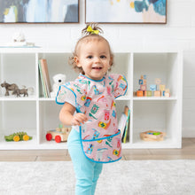 Load image into Gallery viewer, A toddler in a colorful Bumkins Ruffle Apron Bib: Boots and blue pants smiles in a playroom with toy-filled shelves behind.
