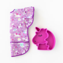 Load image into Gallery viewer, Bumkins Ruffle Apron Bib: Unicorns lays folded lengthwise next to a pink unicorn-shaped divided plate on a white background.

