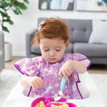 Load image into Gallery viewer, A toddler in a high chair, wearing a Bumkins Ruffle Apron Bib: Unicorns, holds a blue spoon over a pink divided plate of food.
