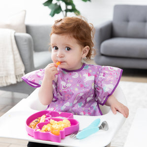 A toddler in a high chair eats snacks from a pink unicorn-shaped plate, wearing a Bumkins Ruffle Apron Bib: Unicorns in a cozy living room.