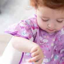 Load image into Gallery viewer, Close-up of a toddler looking down in a high chair, wearing a Bumkins Ruffle Apron Bib: Unicorns.
