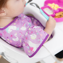 Load image into Gallery viewer, Close-up of a toddler in a high chair holding a blue spoon wearing a Bumkins Ruffle Apron Bib: Unicorns, with focus on the ruffle sleeve.
