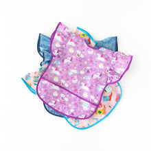 Load image into Gallery viewer, Bumkins Ruffle Apron Bib: Unicorns lays on top of a stack of similar bibs on a white background.
