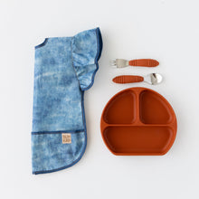 Load image into Gallery viewer, Bumkins Ruffle Apron Bib: Crushed Denim lays folded lengthwise on a white background next to an orange plate and matching utensils.
