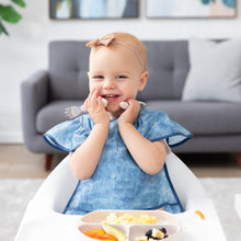 Load image into Gallery viewer, A smiling toddler in a Bumkins Ruffle Apron Bib: Crushed Denim sits in a high chair, holding utensils by their cheeks with a plate nearby.
