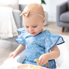 Load image into Gallery viewer, In a bright room, a toddler enjoys food in a high chair, wearing a Bumkins Ruffle Apron Bib: Crushed Denim and a bow headband.
