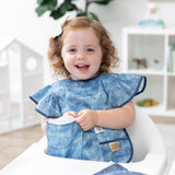A girl wearing Bumkins Ruffle Apron Bib: Crushed Denim smiles in a high chair while opening a matching snack bag.