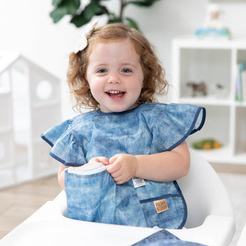 A girl wearing Bumkins Ruffle Apron Bib: Crushed Denim smiles in a high chair while opening a matching snack bag.