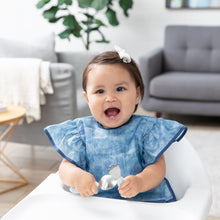 Load image into Gallery viewer, A smiling baby in a bow and a Bumkins Ruffle Apron Bib: Crushed Denim sits in a high chair in a cozy living room, holding utensils.
