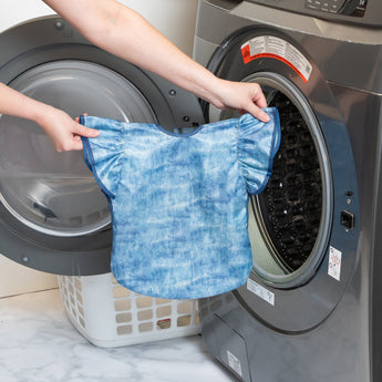 A person holds Bumkins Ruffle Apron Bib: Crushed Denim in front of an open front-loading dishwasher.