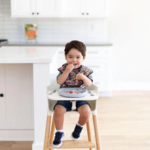 Load image into Gallery viewer, A toddler in a Bumkins SuperBib® 3 Pack: Super Mario™ Power-Up sits in a high chair, eating from a gray divided plate in a bright kitchen.
