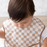 A brown-haired toddler in a tan checkered Bumkins SuperBib® 3 Pack: Palms + Check looks downward.