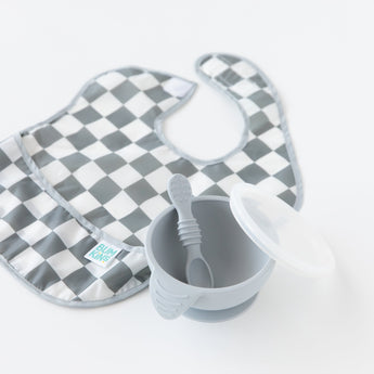 A gray Bumkins SuperBib® 3 Pack: Palms + Check sits gracefully beside a gray bowl with a spoon and lid on a pristine white background.