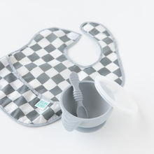 Load image into Gallery viewer, A gray Bumkins SuperBib® 3 Pack: Palms + Check sits gracefully beside a gray bowl with a spoon and lid on a pristine white background.
