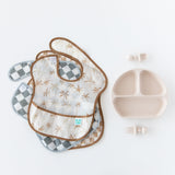 Bumkins SuperBib® 3 Pack: Palms + Check, made from waterproof fabric, next to a divided plate and utensils on a white background.