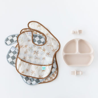 Bumkins SuperBib® 3 Pack: Palms + Check, made from waterproof fabric, next to a divided plate and utensils on a white background.