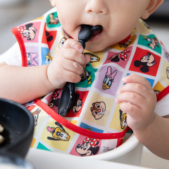 A child in a bright Bumkins SuperBib® 3 Pack: Mickey & Friends MVP holds a spoon, ready to eat from a bowl.