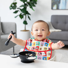 Load image into Gallery viewer, A baby smiles in a colorful Bumkins SuperBib® 3 Pack: Mickey &amp; Friends MVP, holding a spoon near a Mickey Mouse-shaped bowl with yogurt.
