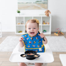 Load image into Gallery viewer, A smiling baby in a high chair, wearing a blue Bumkins SuperBib®: Mickey &amp; Friends MVP, holds a spoon while eating cereal in a bowl.

