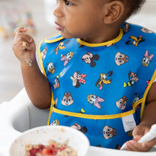 Load image into Gallery viewer, A toddler wears a blue Bumkins SuperBib® 3 Pack: Mickey &amp; Friends MVP, holding cereal, with a bowl of fruit in front.
