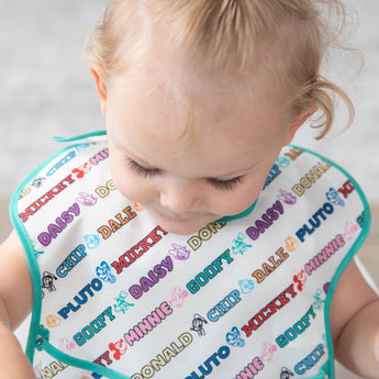 A child wearing a white Bumkins SuperBib® 3 Pack: Mickey & Friends MVP cartoon names like Mickey, Minnie, Pluto & Goofy.