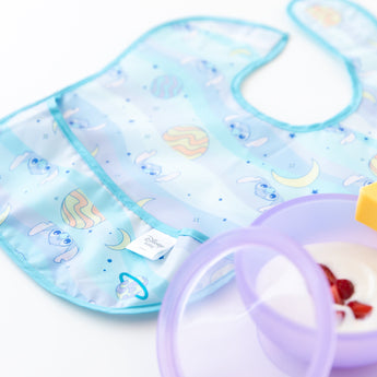 A Bumkins SuperBib® 3 Pack: Stitch Star-ring Up Trouble lays flat next to a purple bowl filled with yogurt and berries.