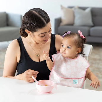 A woman and toddler wearing a Bumkins SuperBib® 3 Pack: Love You Bunches sit at a white table with a pink bowl & spoon.