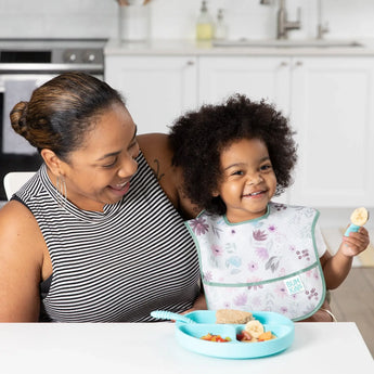 A woman sits with a child in a Bumkins SuperBib® 3 Pack: Love You Bunches, holding a blue fork, at a white table with a blue plate nearby.