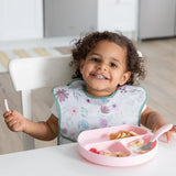 A curly-haired toddler sits at a table, wearing a Bumkins SuperBib® 3 Pack: Love You Bunches and eating from a pink plate with pink utensils.