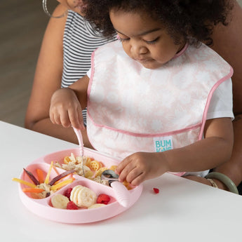 A toddler in a Bumkins SuperBib® 3 Pack: Love You Bunches sits on an adult's lap, eating from a pink plate with a fork and spoon.