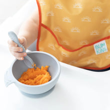 Load image into Gallery viewer, Close-up of a baby in a yellow Bumkins SuperBib® 3 Pack: Desert Boho holding a spoon over orange mash in a bowl on a white highchair tray.
