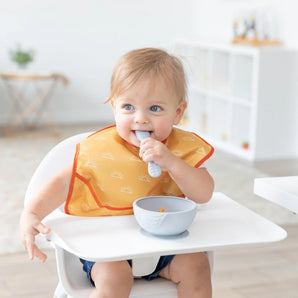 A blonde baby sits in a high chair wearing a yellow Bumkins SuperBib® 3 Pack: Desert Boho, chewing a spoon next to a bowl in a bright room.