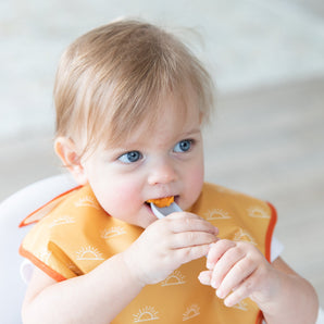 A boy in a yellow bib eats mashed sweet potatoes with a Bumkins Silicone Dipping Spoons 3 Pack: Taffy.