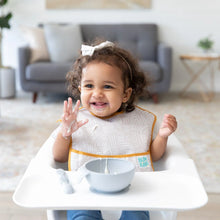 Load image into Gallery viewer, A smiling toddler in a high chair, wearing a beige Bumkins SuperBib® 3 Pack: Desert Boho, plays with food from a gray bowl.
