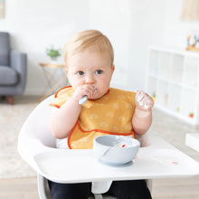 Load image into Gallery viewer, In a bright room, a baby in a high chair wearing a yellow Bumkins SuperBib® 3 Pack: Desert Boho eats from a bowl while holding a spoon.

