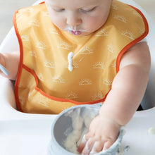Load image into Gallery viewer, A baby in high chair wears a yellow Bumkins SuperBib® 3 Pack: Desert Boho, dripping yogurt down the front as they eat from a bowl.
