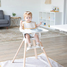 Load image into Gallery viewer, A baby in a high chair, wearing a beige Bumkins SuperBib® 3 Pack: Desert Boho, eats from a gray plate with utensils in a bright living room.
