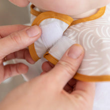 Load image into Gallery viewer, Close-up of hands fastening the hook-and loop closure of a beige Bumkins SuperBib® 3 Pack: Desert Boho around a baby's neck.
