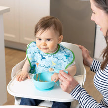 Load image into Gallery viewer, A baby in a Bumkins SuperBib® 3 Pack: Rolling With the Waves sits in a high chair beside a smiling woman, with food in a bowl on the tray.
