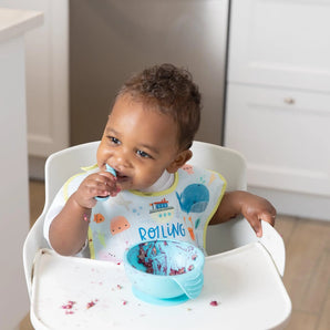 A toddler wearing a Bumkins SuperBib® 3 Pack: Rolling With the Waves in a high chair messily eats from a blue bowl with a spoon.