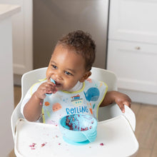 Load image into Gallery viewer, A toddler wearing a Bumkins SuperBib® 3 Pack: Rolling With the Waves in a high chair messily eats from a blue bowl with a spoon.
