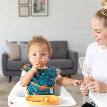 Load image into Gallery viewer, A toddler in a Bumkins SuperBib® 3 Pack: All Together Now eats in a high chair while a smiling woman sits nearby in a cozy living room.
