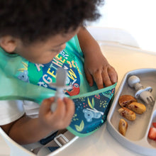 Load image into Gallery viewer, A toddler in a high chair wearing a Bumkins SuperBib® 3 Pack: All Together Now holds a gray spoon while reaching into the bib's pocket.
